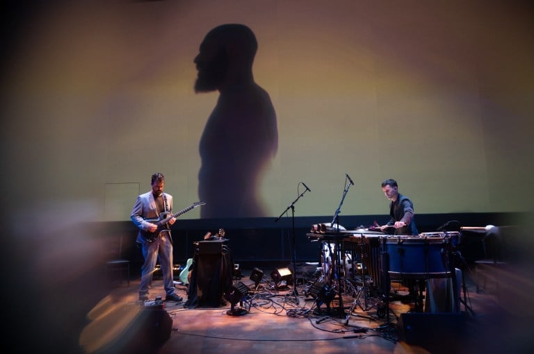 Two musicians playing on stage. A man's shadow is projected onto the wall.