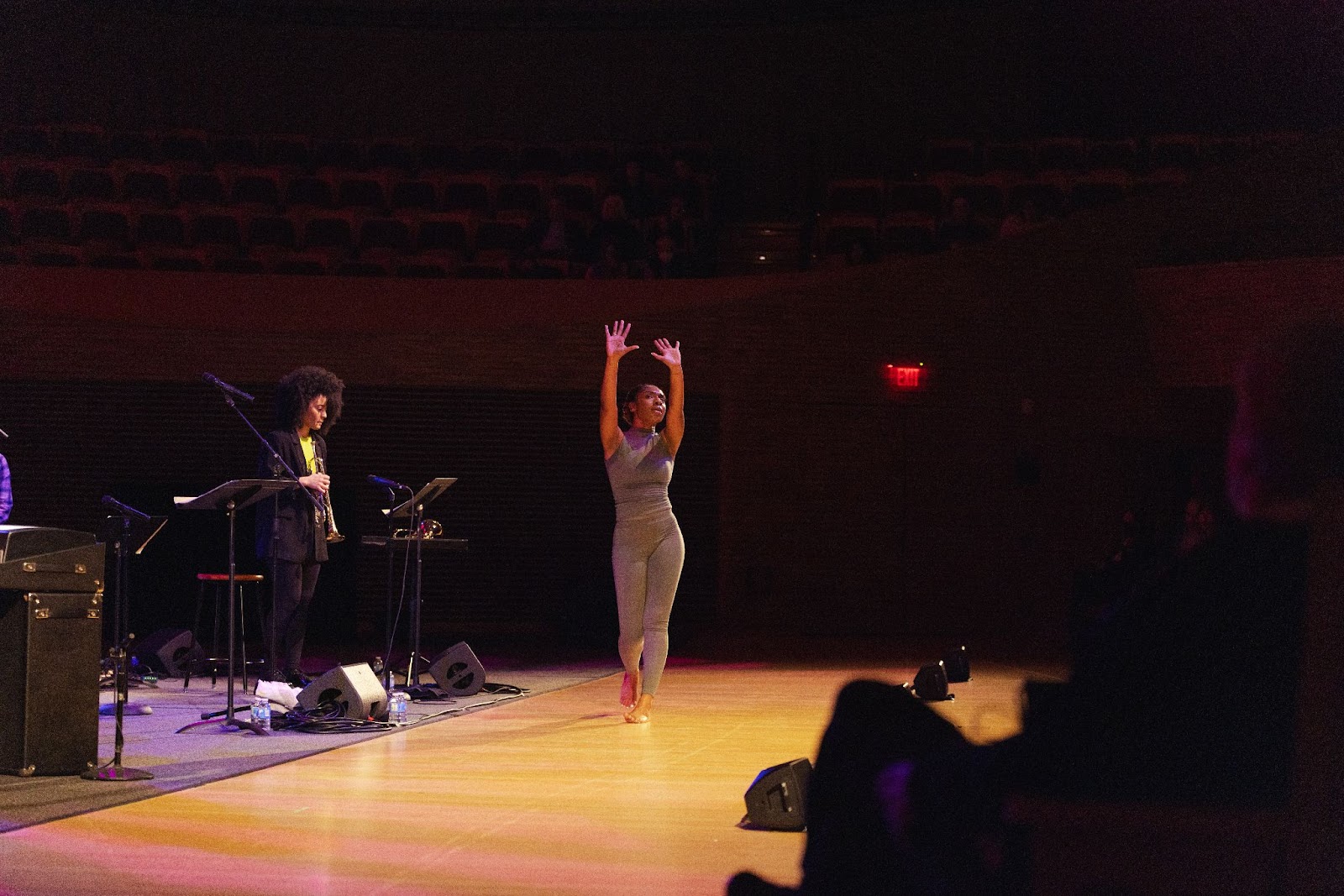 A dancer in a silver two-piece raises her hands to the ceiling as people perform in the back.