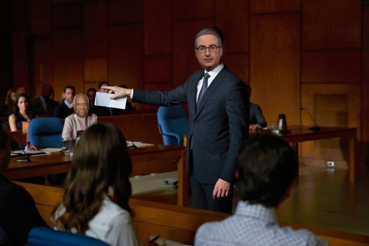 A courtoom with a lawyer pointing, whose face has been photoshopped with John Oliver's, pointing to a man on the stands whose face has been photoshopped with Justice Clarence Thomas'.