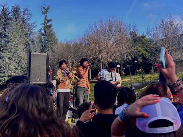 Six of Spades band member perform on a stage in front of Terman Fountain and a crowd of students.