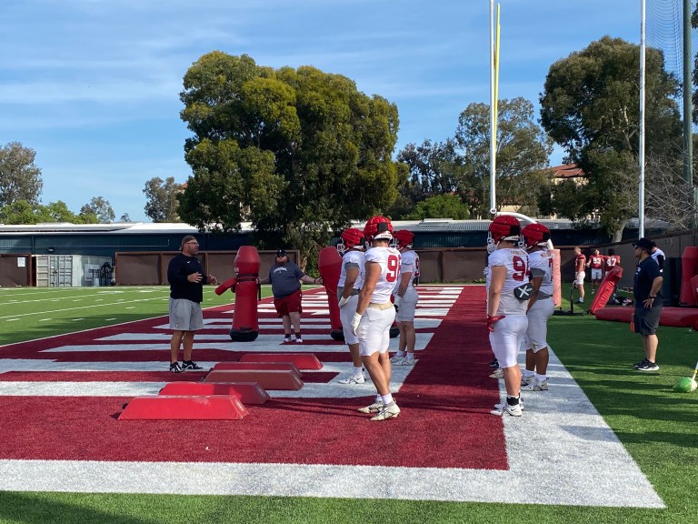 Players in red and white Stanford uniforms stand in the end zone looking towards a coach.