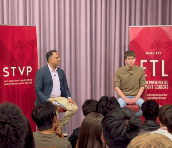 Two men sit on stools in front of a crowd.