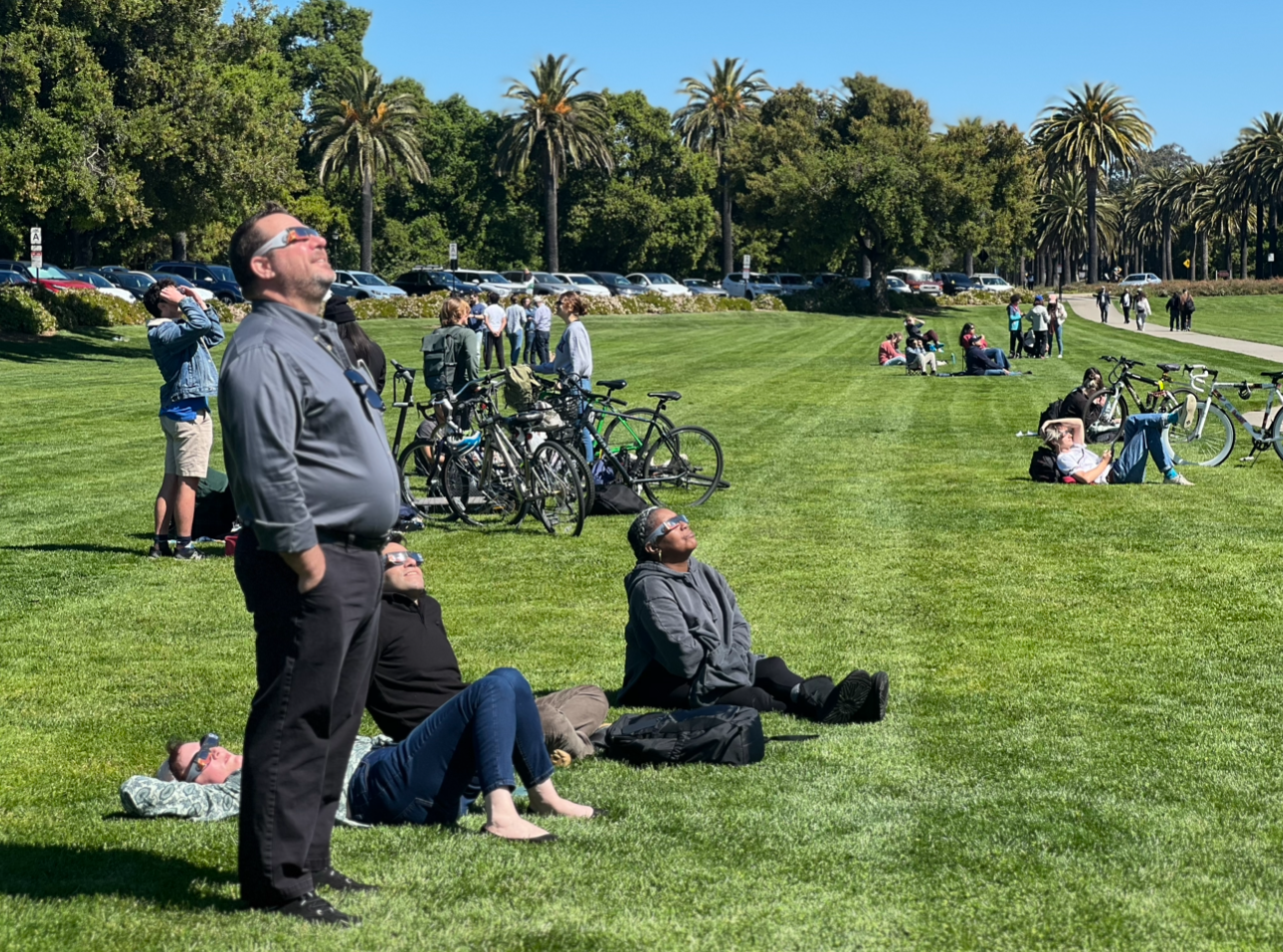 Several people sitting and standing on the Oval, wearing solar glasses and looking up at the eclipse.