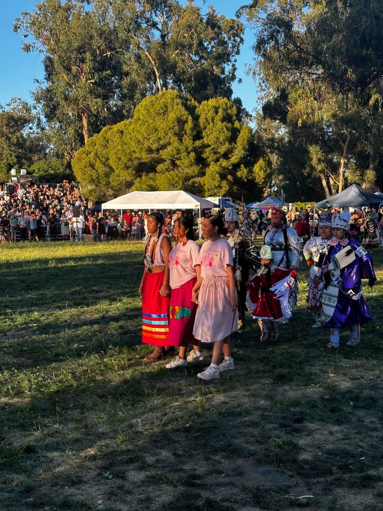 Three female students wearing bright pink "Powwow" T-shirts walk hand-in-hand in a grass field. Women dressed in Native American traditional dress, called regalia, walk behind them.