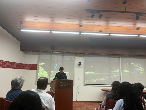 A speaker is talking in a room of audience with "Stanford Humanities center" written across the walls