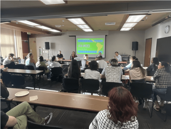 Photo of people sitting behind desks in front of a projector and four speakers.