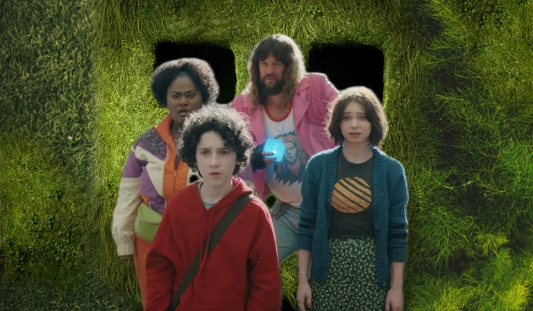 Four individuals stand in front of a green, moss-covered wall with two dark rectangular openings. There is a boy, a girl, a woman, and a man holding a glowing cube.