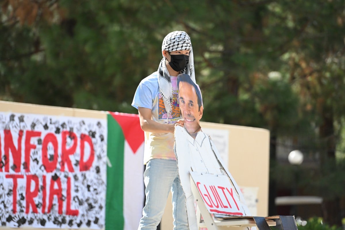 A protester wearing a keffiyeh and a face mask holds a cardboard cutout of University president Jonathan Levin.