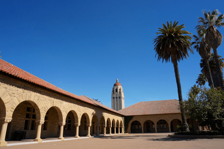 Picture of sandstone archways of the main quadrangle of campus with Hoover Tower in the background, palm trees on the side.