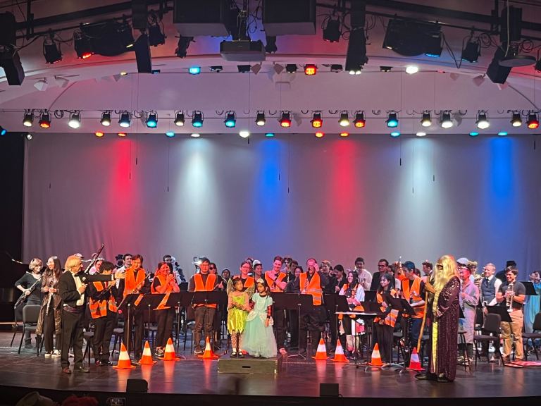 members of The Stanford Symphony Orchestra and Wind Symphony stand on stage wearing bright orange construction vests amongst other costumes