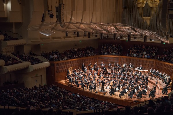 A birds eye view of The San Francisco Symphony sitting at their respective music stands on a large stage